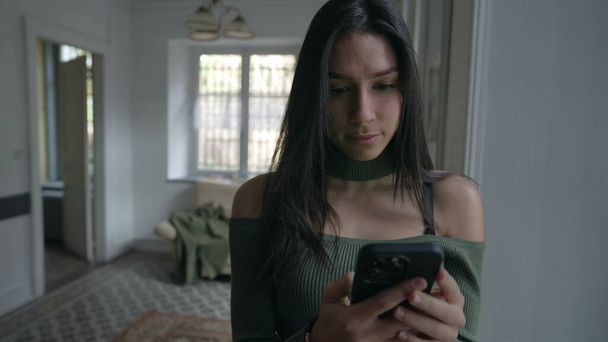 Jedna mladá žena drží telefon a dívá se na obrazovku, zatímco stojí doma u okna. Žena používající moderní technologie - Fotografie, Obrázek