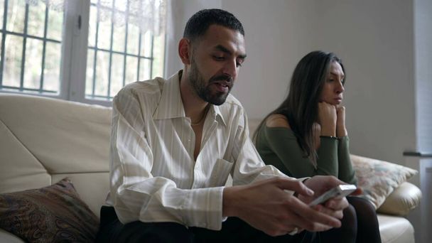 Ένα ζευγάρι σε κρίση. Αγνοεί την κοπέλα του ενώ κρατάει κινητό στο σπίτι. Φιλενάδα θέλει προσοχή από συνεργάτη απορροφάται από το περιεχόμενο των μέσων κοινωνικής δικτύωσης σε απευθείας σύνδεση κάθεται στον καναπέ σε εσωτερικούς χώρους - Φωτογραφία, εικόνα