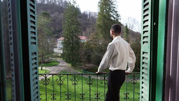 Πίσω από ένα στοχαστικό νεαρό άνδρα που στέκεται έξω στο μπαλκόνι του σπιτιού κοιτάζοντας τον κήπο. Πολυτελής τρόπος ζωής ενός σκεπτικού προσώπου - Φωτογραφία, εικόνα
