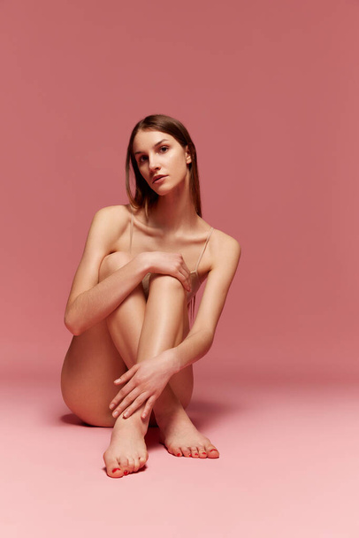 Portrait of tender, young girl with slim healthy body, sitting on floor, posing in beige underwear against pink studio background. Concept of beauty, body care, fitness, sport, health, figure - Φωτογραφία, εικόνα