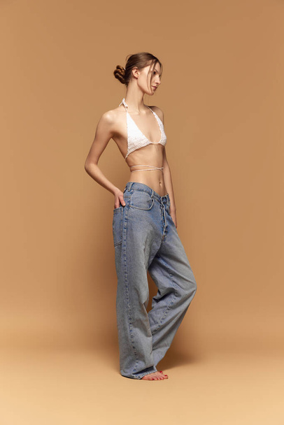 Portrait of young girl with slim body posing in oversized jeans and knitted top against light brown studio background. Concept of beauty, body care, fitness, sport, health, figure, fashion - 写真・画像
