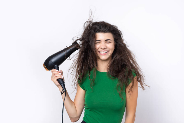 A cute young girl dressed in a green top dries her beautiful long silky hair with a hair dryer against a white background. - Foto, Imagen
