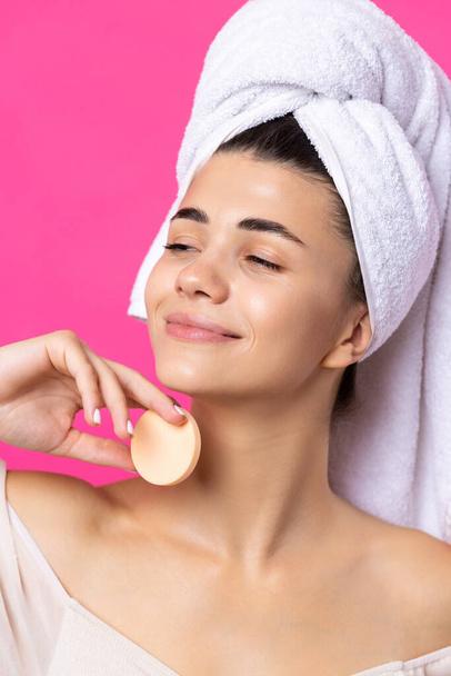 Beautiful cheerful attractive girl with a towel on her head, holds a sponge against a pink background. - Photo, image