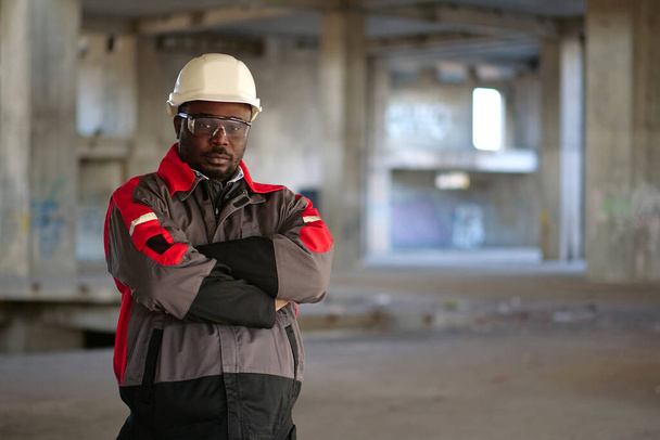 Αφροαμερικάνος εργάτης με λευκό σκληρό καπέλο και προστατευτικά γυαλιά στέκεται στο εργοτάξιο και κοιτάζει την κάμερα - Φωτογραφία, εικόνα