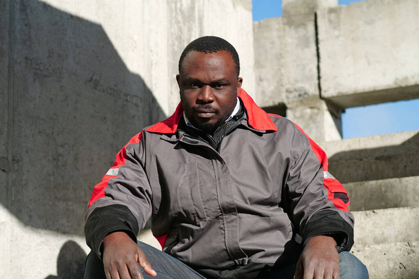 Afrikkalainen amerikkalainen työntekijä istuu portaissa rakennusalueella ja katsoo kameraa - Valokuva, kuva