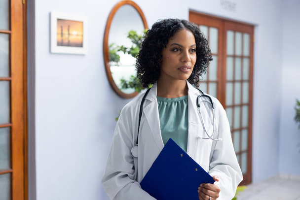 Biracial female doctor wearing stethoscope and holding clipboard at doctor's office. Hospital, medicine and healthcare. - Foto, Bild