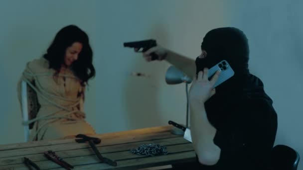 Un kidnappeur parle sur son portable et menace une femme dans un immeuble abandonné. Un homme en cagoule noire avec une arme. Concept de kidnapping. Mouvement lent - Séquence, vidéo