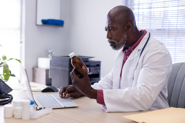African american male doctor sitting at desk holding medicine and using laptop at doctor's office. Hospital, medicine, healthcare and communication. - Фото, зображення