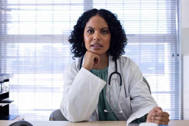 Biracial female doctor sitting at desk and having video call at doctor's office. Hospital, medicine, healthcare and communication. - Foto, Imagem