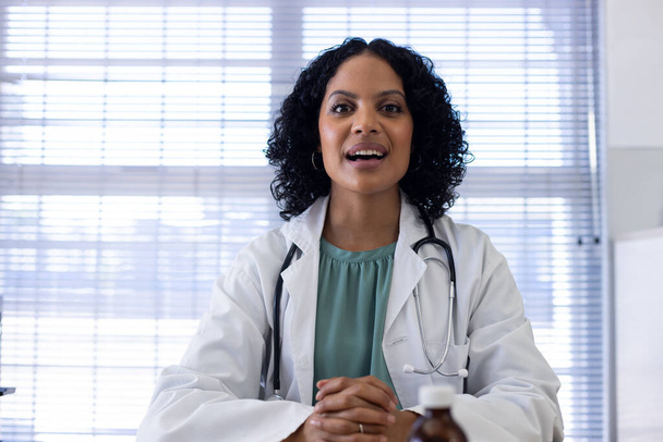 Biracial female doctor sitting at desk and having video call at doctor's office. Hospital, medicine, healthcare and communication. - Foto, Bild