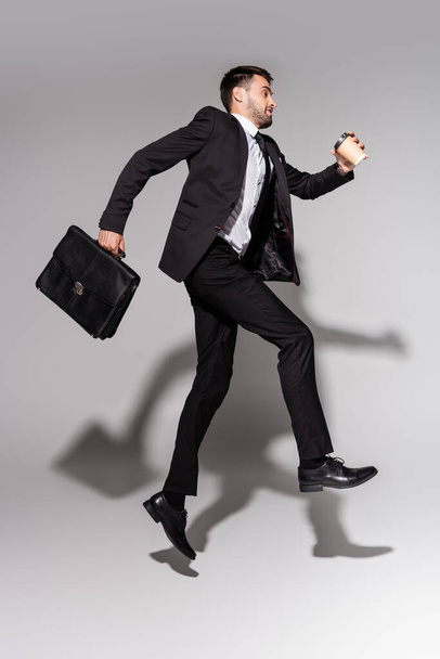 side view of stylish businessman in black suit levitating with coffee to go and leather briefcase on grey background - Foto, Imagem