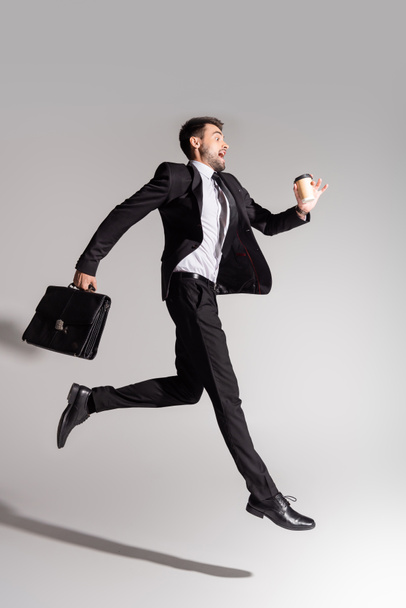 side view of thrilled businessman in black suit levitating with leather briefcase and paper cup on grey background - Photo, Image