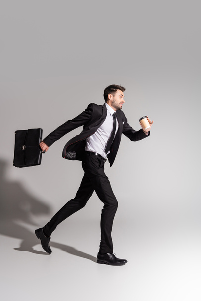 side view of positive businessman in black suit running with briefcase and paper cup on grey background - Foto, Imagem