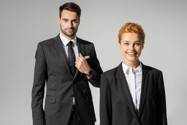 cheerful redhead businesswoman smiling at camera near confident businessman in stylish suit isolated on grey - 写真・画像
