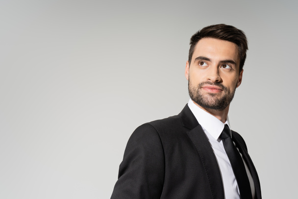 smiling and dreamy businessman in black blazer looking away isolated on grey - Foto, Imagem