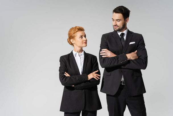 positieve business collega 's in zwarte blazers staan met gevouwen armen en kijken naar elkaar geïsoleerd op grijs - Foto, afbeelding