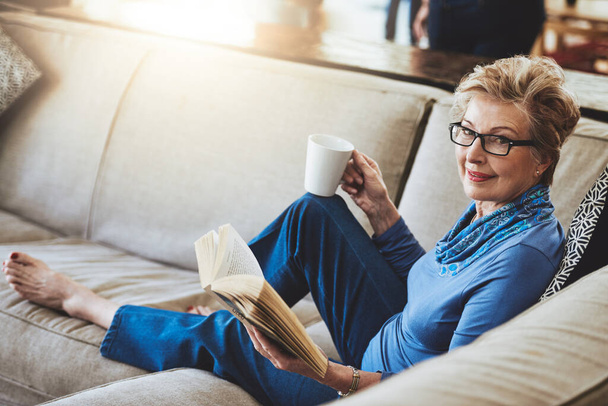 The retired life aka the good life. Portrait of a senior woman relaxing with a warm beverage and a book on the sofa at home - Foto, Imagem