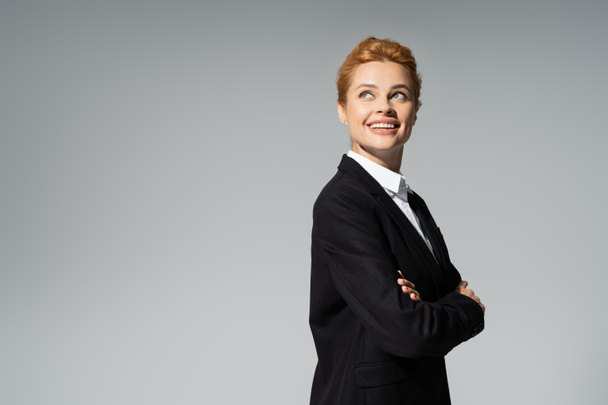 happy redhead businesswoman in black blazer looking away while standing with crossed arms isolated on grey - Foto, Imagem