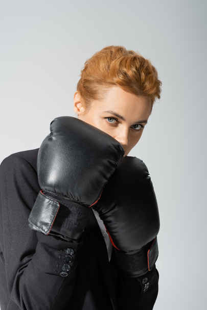 redhead businesswoman obscuring face with boxing gloves while looking at camera isolated on grey - 写真・画像