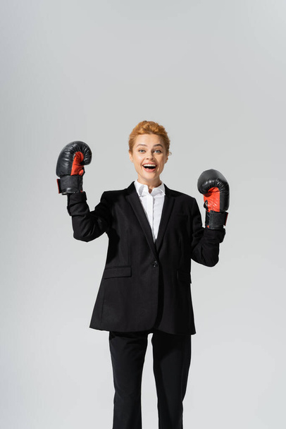 opgewonden roodharige zakenvrouw in zwart pak en latex handschoenen vreugde geïsoleerd op grijs - Foto, afbeelding