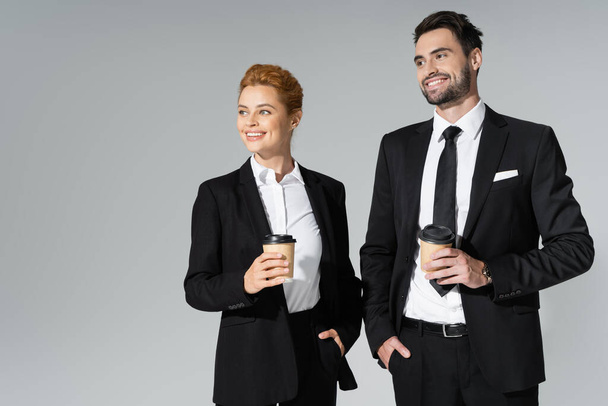 radośni partnerzy biznesowi trzymający kawę na wynos, stojący z rękami w kieszeniach i patrzący odizolowani na szaro - Zdjęcie, obraz