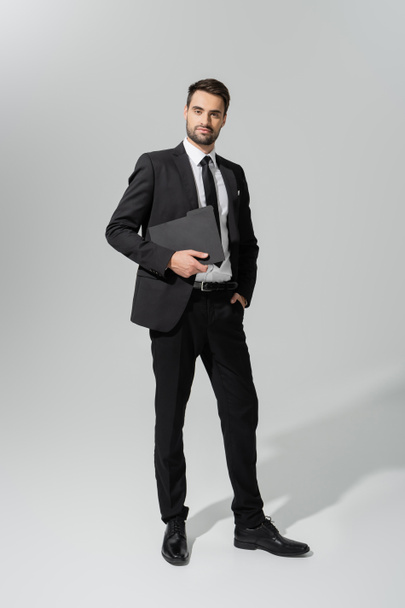  longitud completa de empresario exitoso y seguro en traje elegante negro de pie con carpeta sobre fondo gris - Foto, Imagen