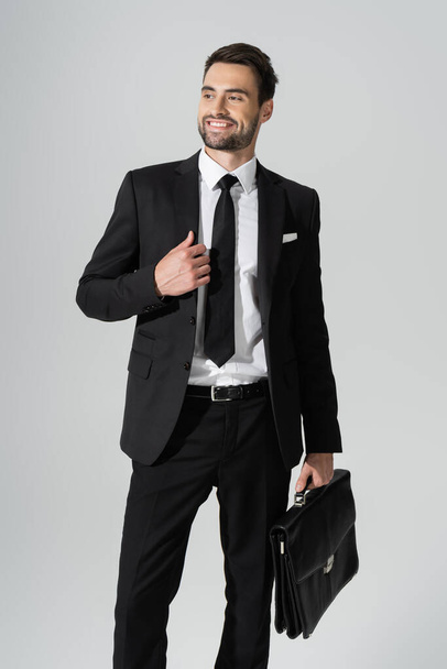fröhlicher Geschäftsmann in schwarzem, stylischem Hosenanzug, der mit Aktentasche dasteht und isoliert auf grau blickt - Foto, Bild