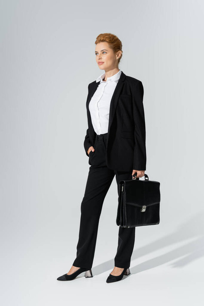 full length of positive businesswoman in black pantsuit posing with briefcase and hand in pocket on grey background - Fotó, kép