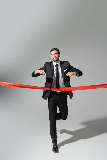 full length of happy businessman in black suit running and crossing red ribbon on grey background - Photo, Image