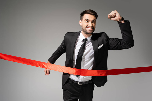 exitoso y emocionado hombre de negocios mostrando gesto de victoria y cruzando la cinta de acabado rojo aislado en gris - Foto, Imagen