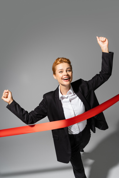 happy businesswoman showing win gesture while crossing finish ribbon on grey background - Photo, Image