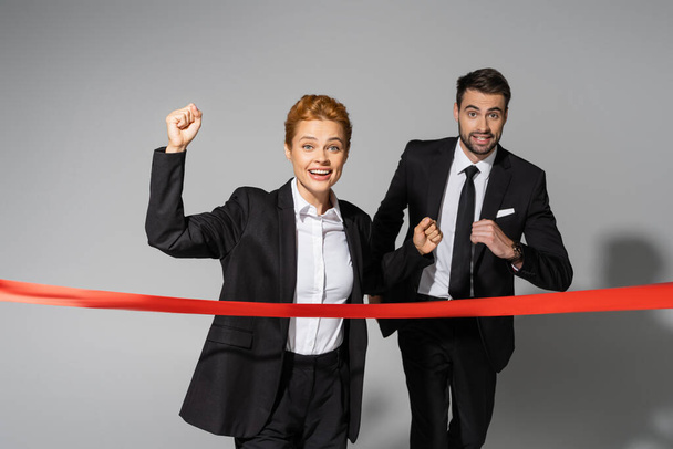 cheerful redhead businesswoman showing win gesture near finish ribbon and business competitor running on grey background - Photo, Image