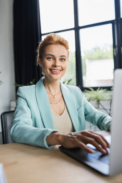 успешная деловая женщина в стильном блейзере, печатающая на ноутбуке и улыбающаяся на камеру в офисе - Фото, изображение