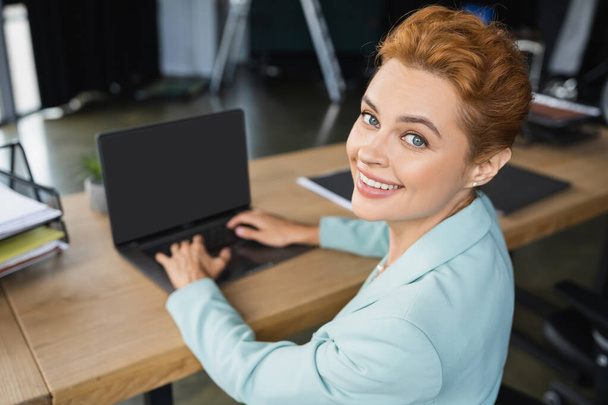 alegre pelirroja mujer de negocios escribiendo en borrosa portátil con pantalla en blanco y sonriendo a la cámara en la oficina - Foto, Imagen