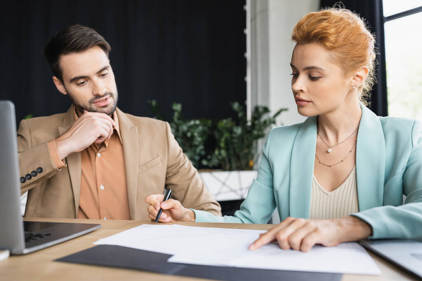 redhead businesswoman pointing at documents near thoughtful colleague in office - Φωτογραφία, εικόνα