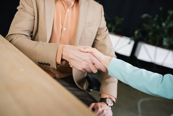 partial view of business partners shaking hands on blurred foreground in office - Photo, Image