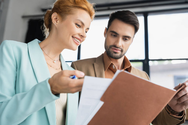cheerful redhead businesswoman showing documents to bearded colleague in office - Фото, зображення