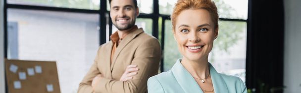 successful redhead businesswoman looking at camera near colleague smiling on blurred background in office, banner - Фото, изображение