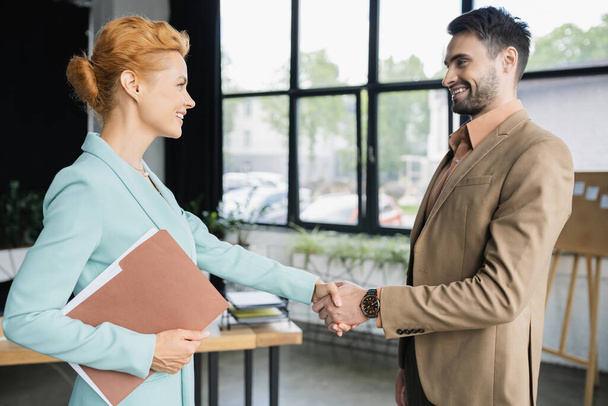 side view of pleased businesswoman with folder shaking hands with young partner in office - Photo, Image