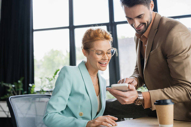 gestionnaire positif montrant téléphone portable à la femme d'affaires dans les lunettes dans le bureau - Photo, image