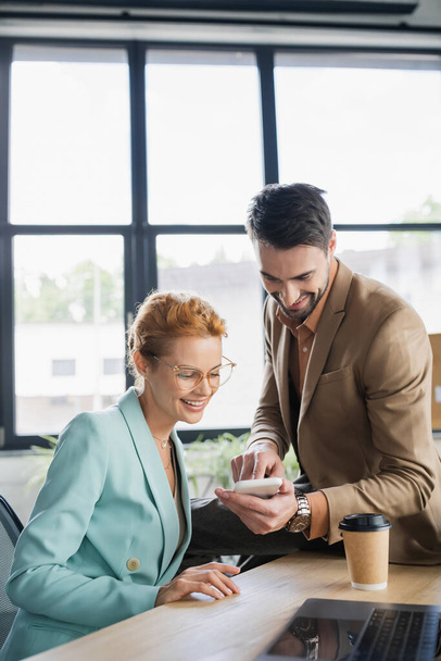 smiling bearded businessman showing smartphone to woman in eyeglasses in office - Fotó, kép