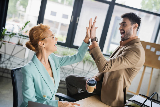 cheerful man with coffee to go giving high five to businesswoman sitting at workplace in office - 写真・画像