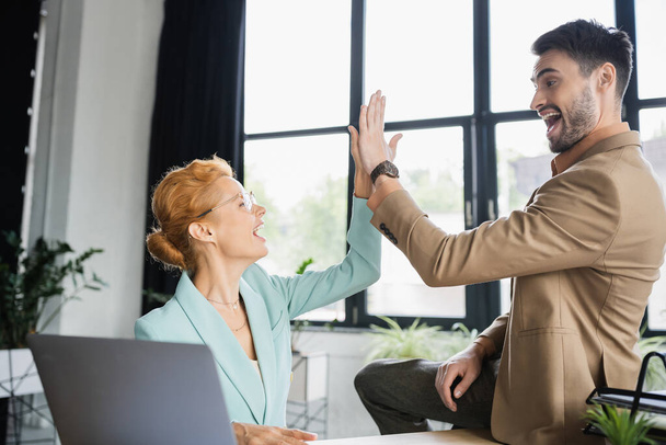 overjoyed business colleagues giving high five near blurred laptop in office - Foto, afbeelding