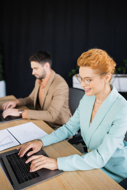 redhead businesswoman in eyeglasses typing on laptop near blurred colleague in office - Foto, afbeelding