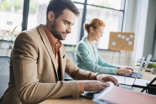 positive bearded businessman typing on laptop near blurred colleague in office - Фото, зображення