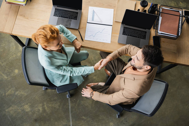 Обзор бизнес-партнеров, пожимающих руки возле ноутбуков и документов с таблицами в офисе - Фото, изображение