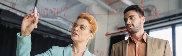 деловая женщина с войлочной ручкой на стеклянной доске рядом с бородатым коллегой в офисе, баннер - Фото, изображение