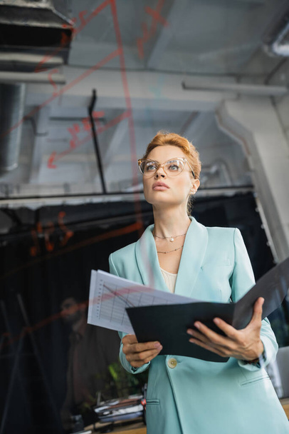 visão de baixo ângulo de empresária ruiva em óculos segurando documentos e olhando para sinais em placa de vidro no escritório - Foto, Imagem