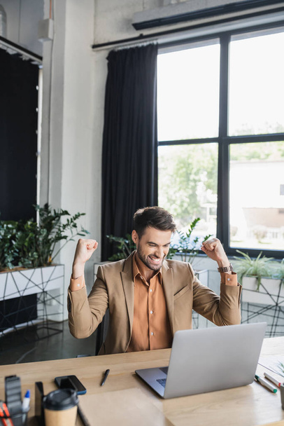 vzrušený podnikatel ukazuje triumfální gesto, zatímco sedí v blízkosti notebooku v kanceláři - Fotografie, Obrázek