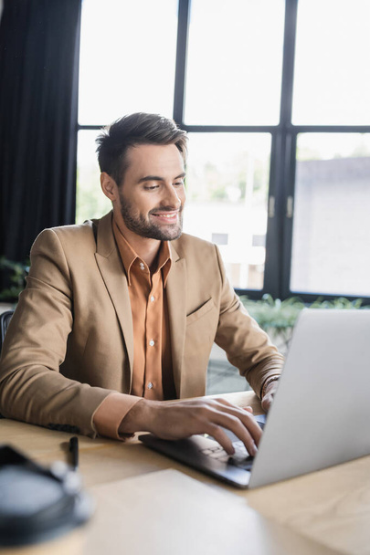 smiling businessman in stylish blazer typing on laptop while working in modern office - Foto, Imagem